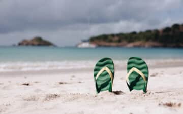 Chinelos verdes enterrados numa praia vazia com mar ao fundo, simbolizando férias